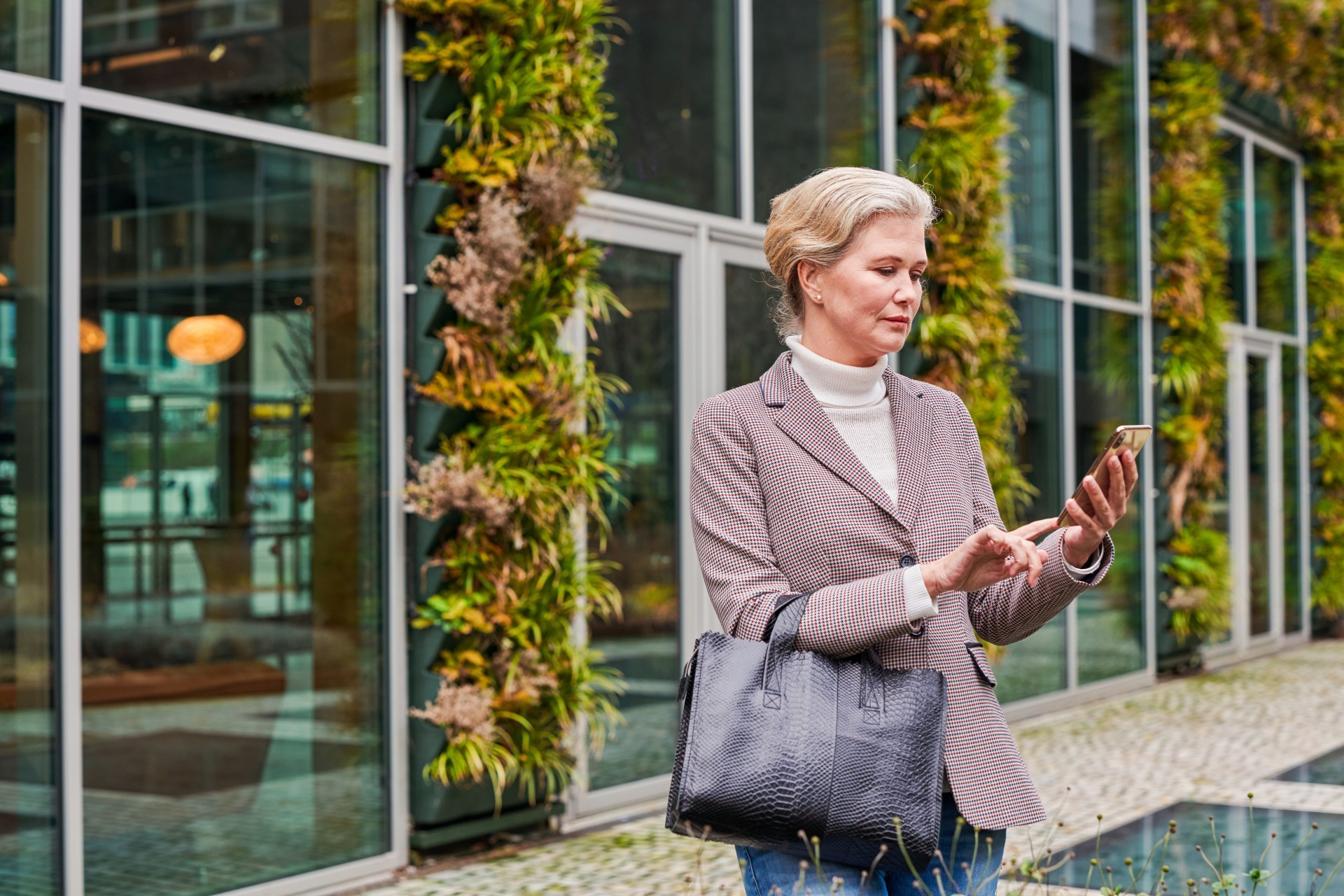 Net geklede vrouw staat voor bedrijfspand waar planten overheen groeien en kijkt naar haar telefoon