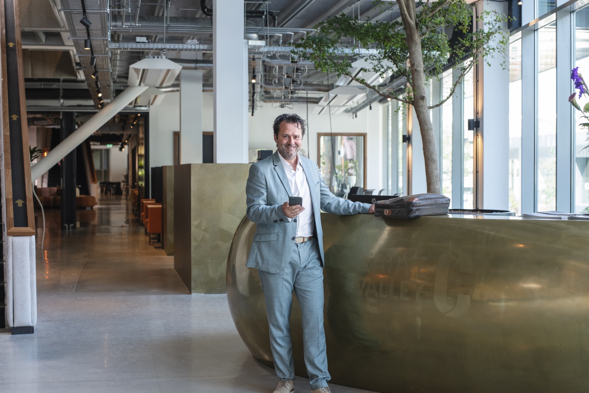 Man in pak staat trots naast een enorme bloempot met boom in de hal van zijn bedrijfspand