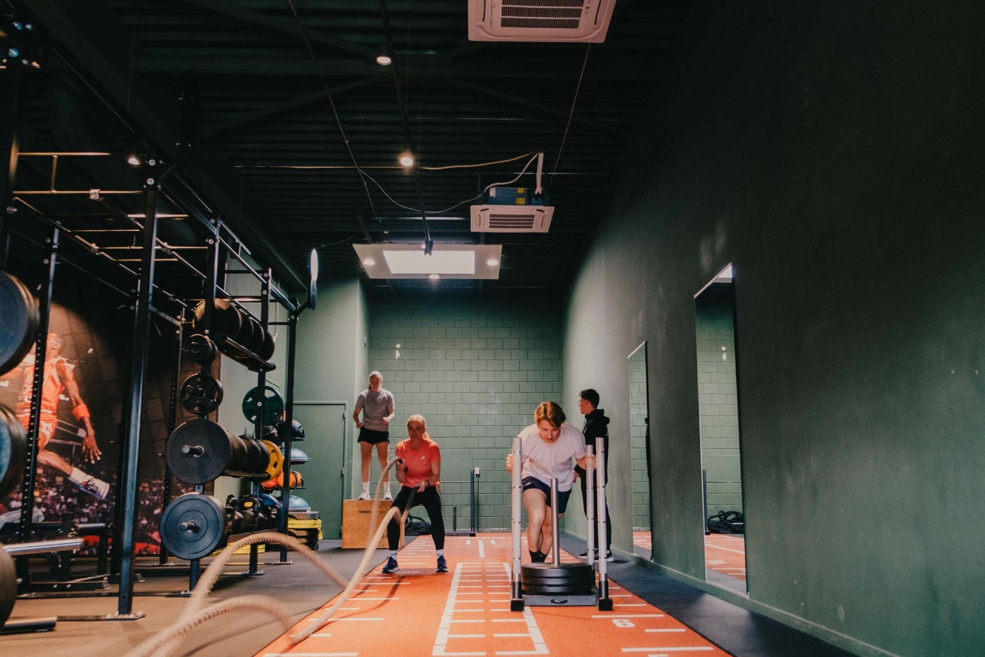 Sporters zijn aan de slag in de sportruimte van Sport Medisch Centrum Amersfoort, waaronder het voortduwen van een prowler, het gebruiken van battle ropes en het uitvoeren van een box step-up oefening