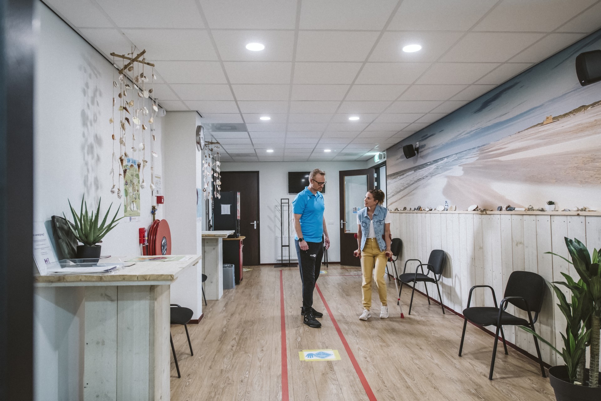 Vrouw op krukken praat met behandelaar in de wachtkamer van een fysiopraktijk