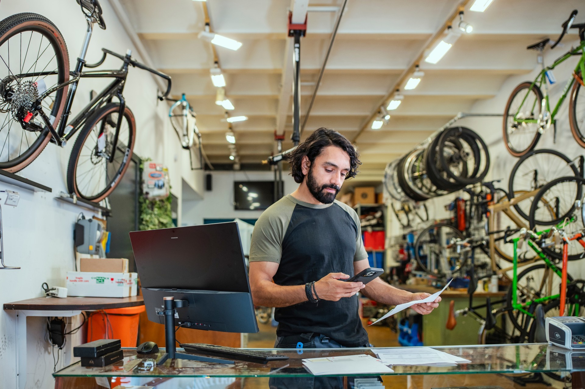 Eigenaar staat in zijn fietsenwinkel achter zijn bureau met computer. In zijn hand heeft hij zijn telefoon terwijl hij papieren controleert.